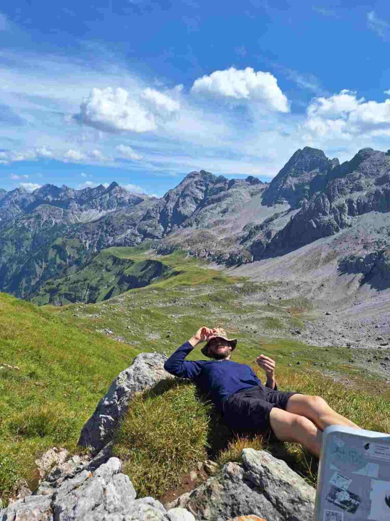 Sportler entspannt sich beim Wandern in den Alpen auf einer Wiese; Symbolbild für aktive Erholung, Stressabbau und die Wichtigkeit von Pausen für die körperliche Leistungsfähigkeit. Feel Good Coaching.